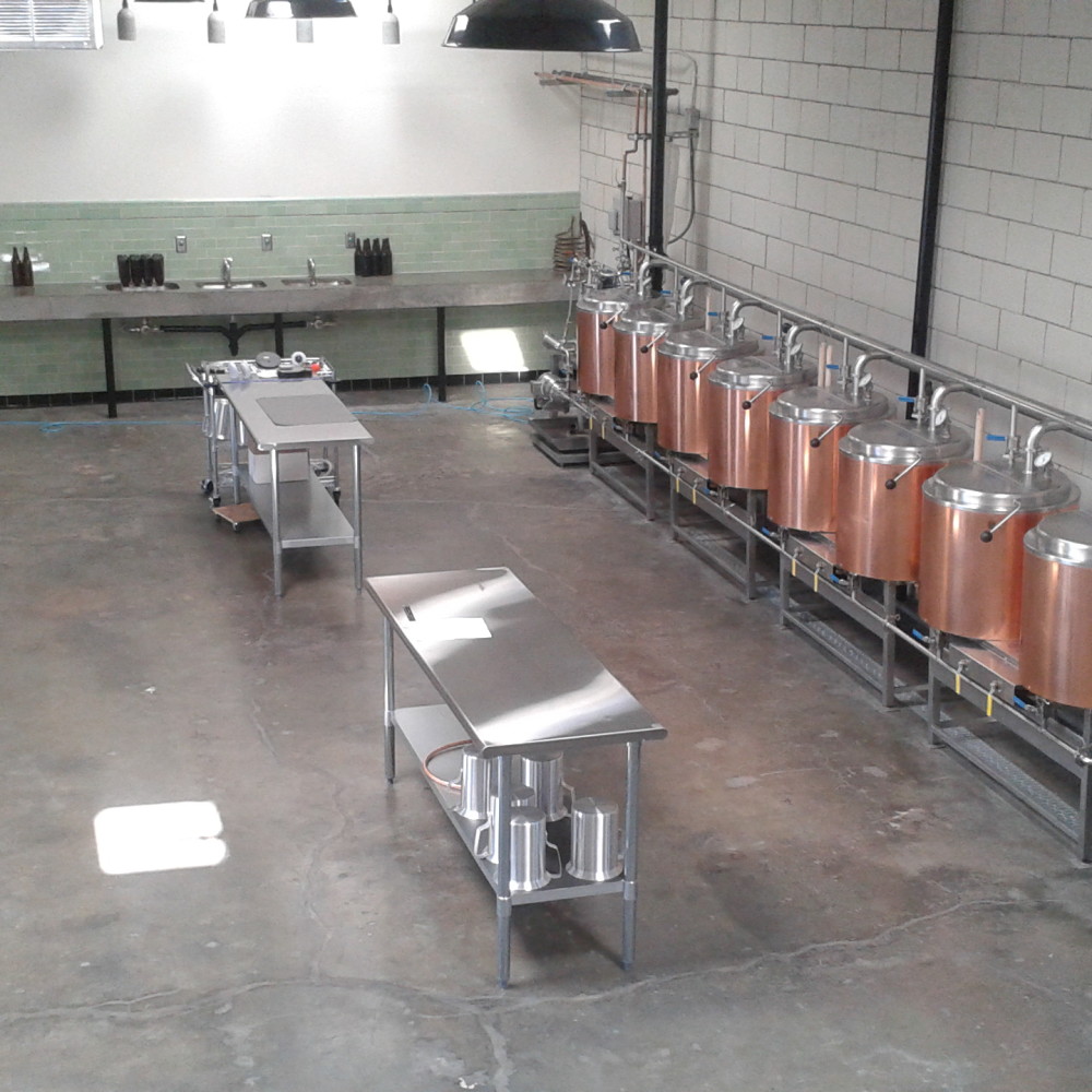 Image is an aerial view of the brewing area, with metal tables in the middle and the copper kettles on the right